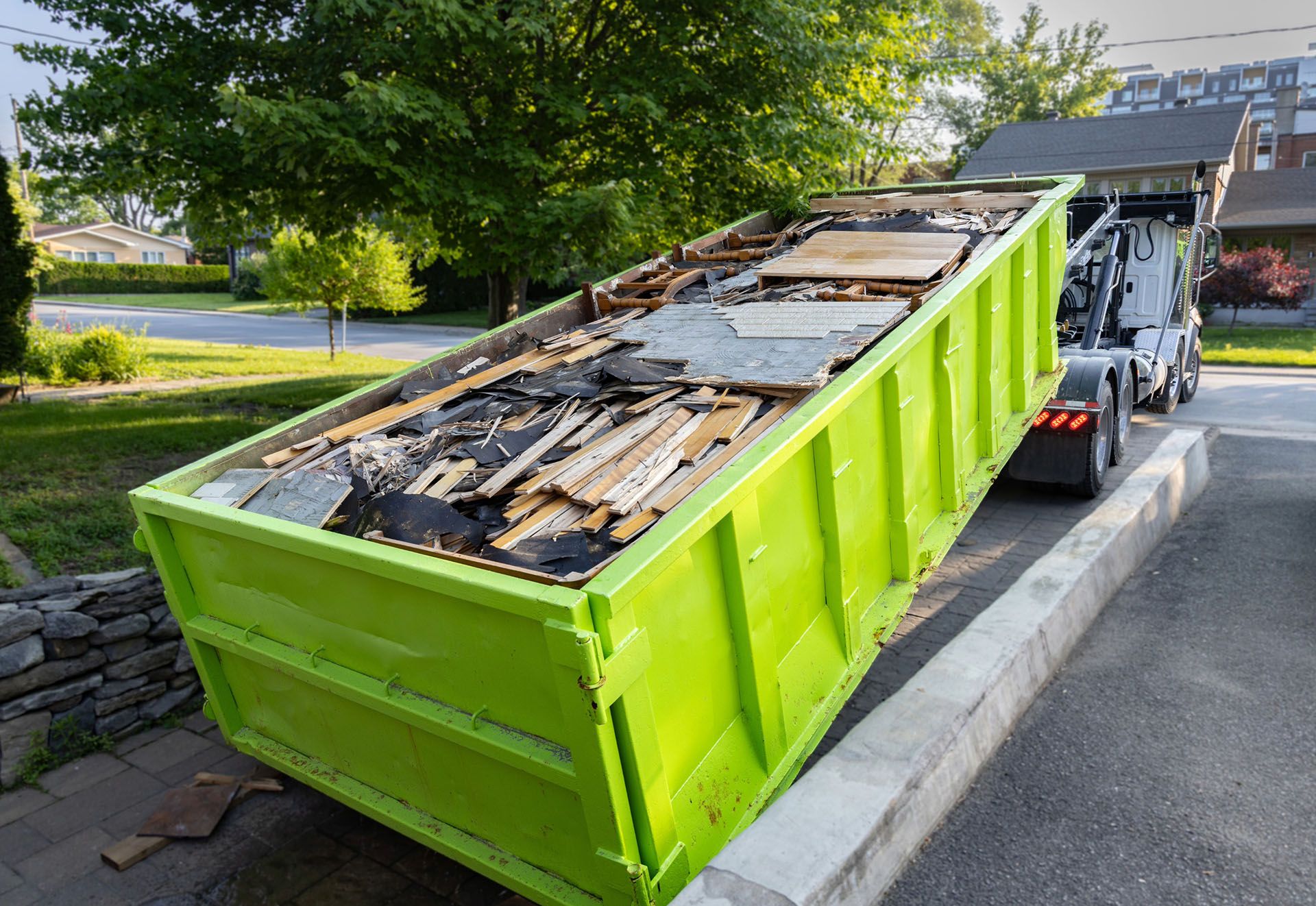 Lime green dumpster overflowing with debris being lifted by a truck in a residential driveway.
