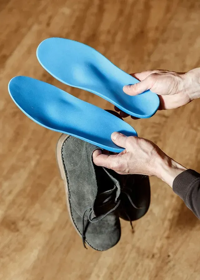 Hands holding two blue orthotic insoles above a pair of gray shoes on a wooden floor.