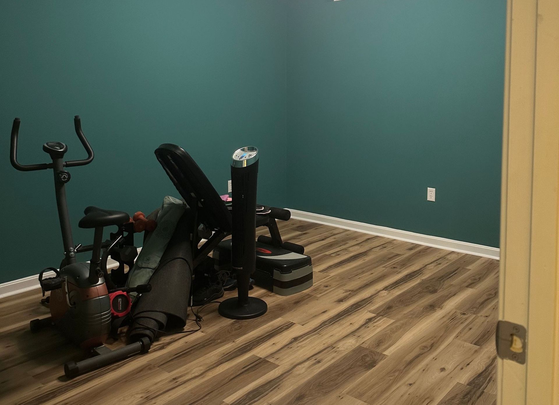 A home gym with teal walls, wood-look floor, and exercise equipment. An upright exercise bike and weight bench are visible.