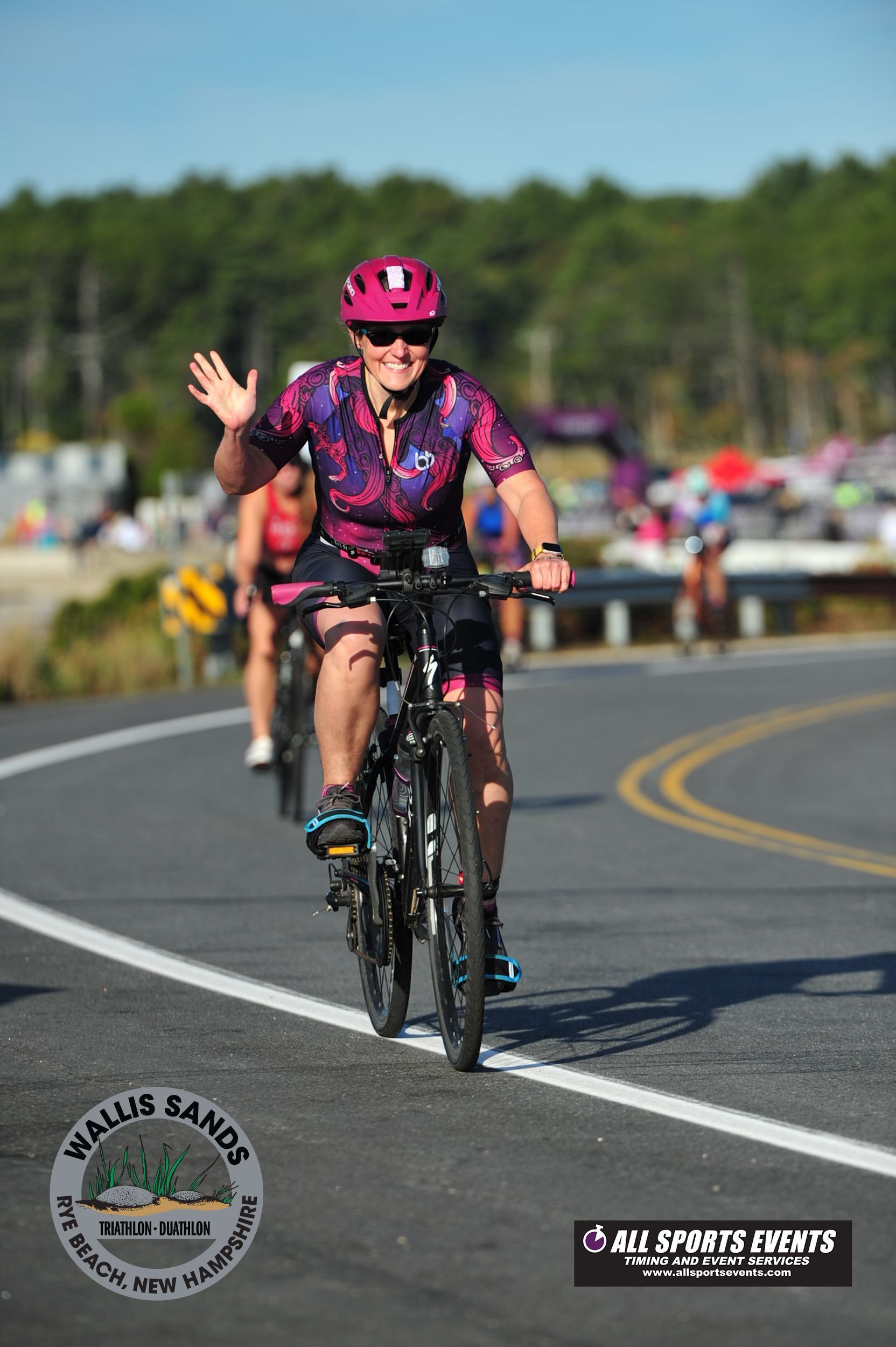 Cyclist in pink helmet waving while riding bike on road, surrounded by others; ocean and trees in background.