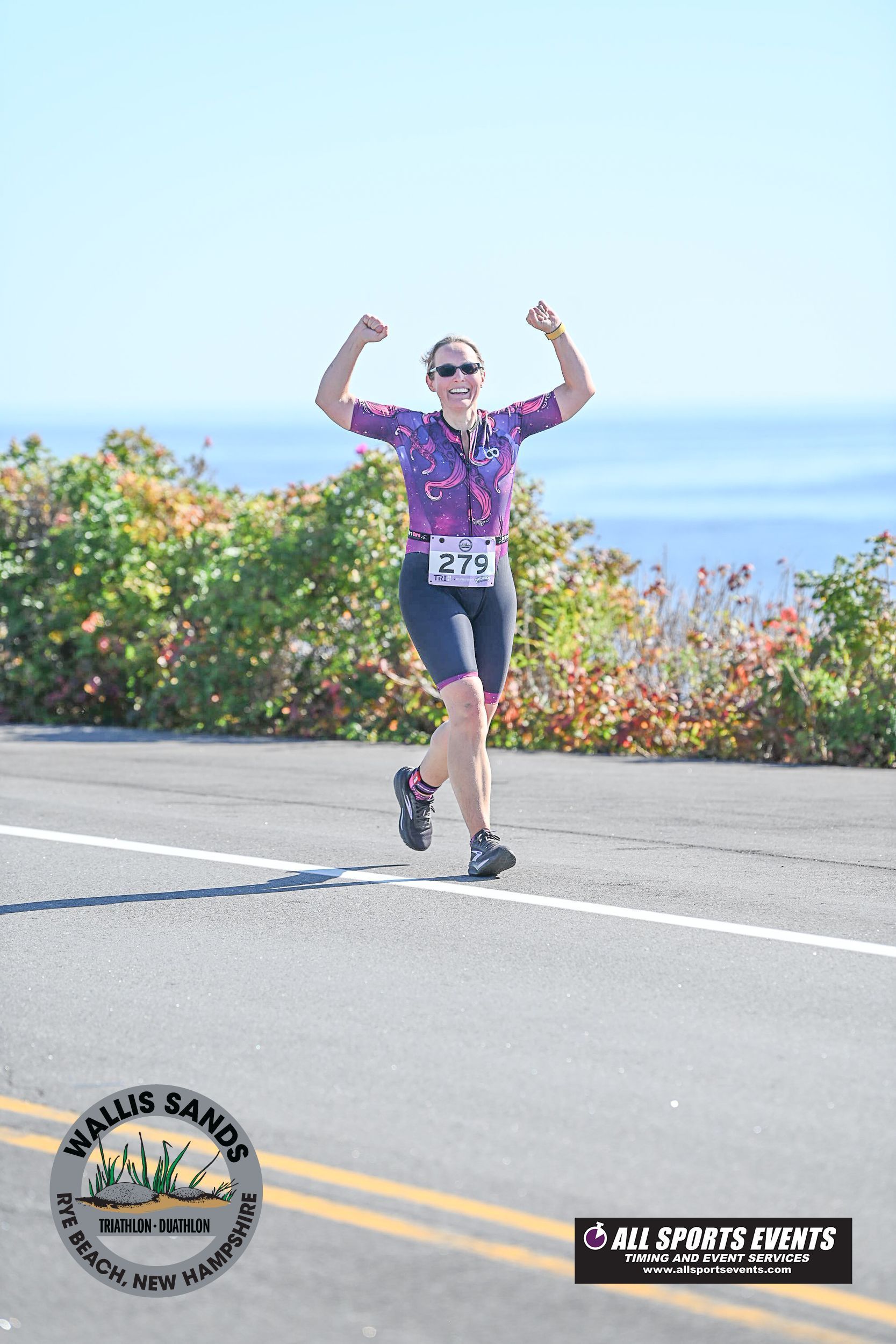 Runner with arms raised on a road near ocean, wearing race bib 