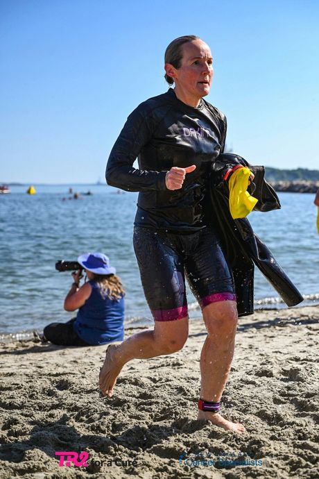 Runner in black wetsuit carrying fins, jogging along a sandy beach near the water.