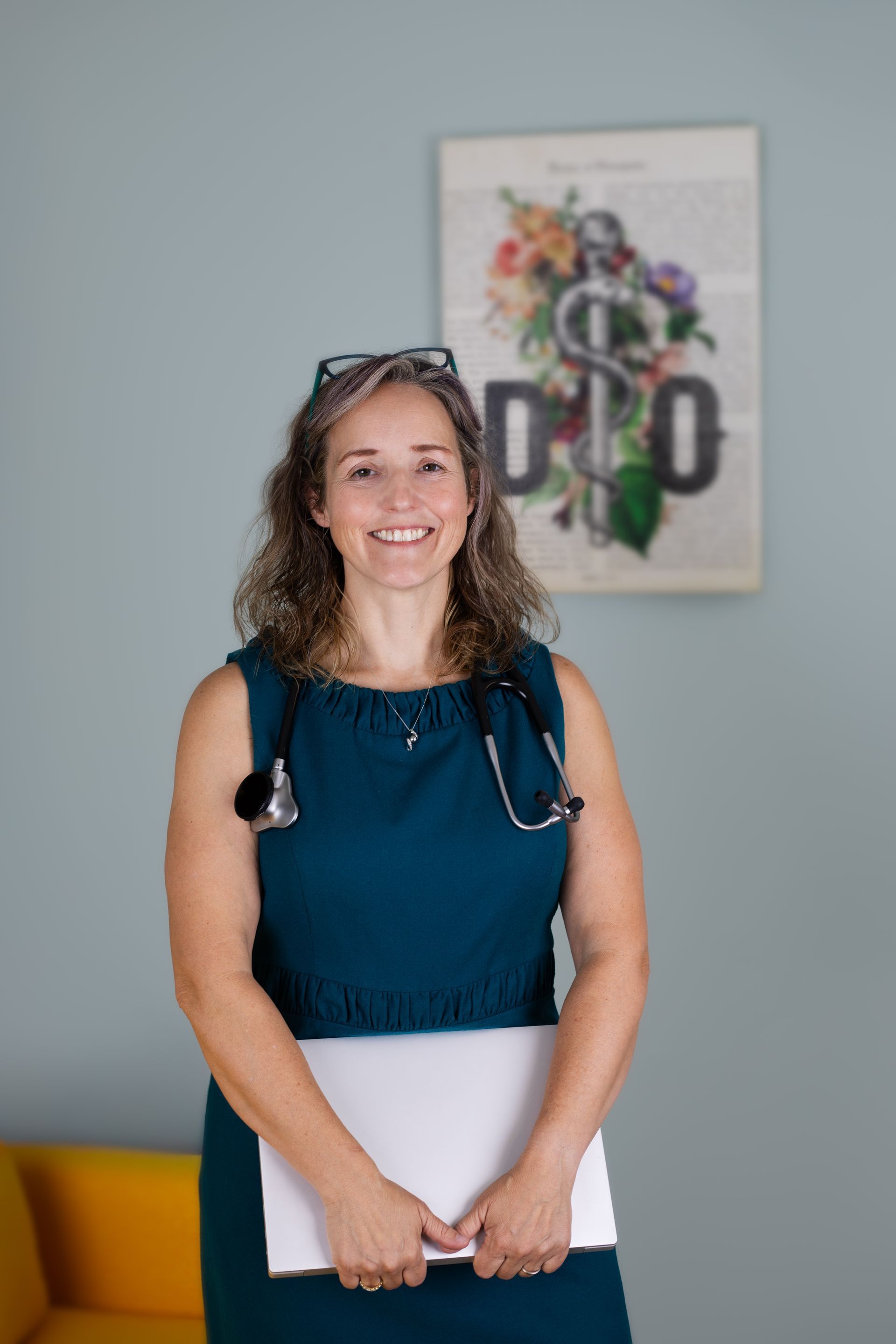 Woman doctor smiling, holding a laptop, wearing a stethoscope, in a teal dress, in an office.