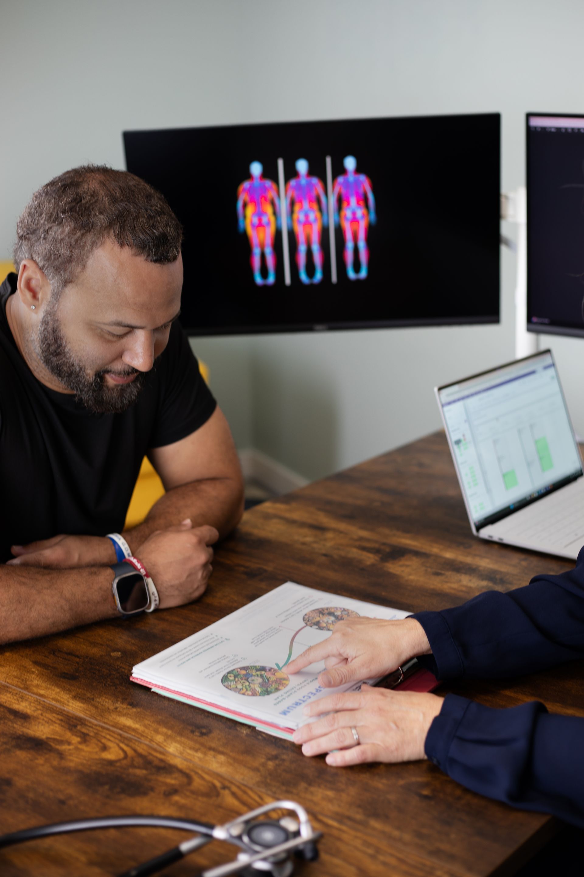 Man looks at document, being reviewed by person at table. TV behind shows body temperature scans.