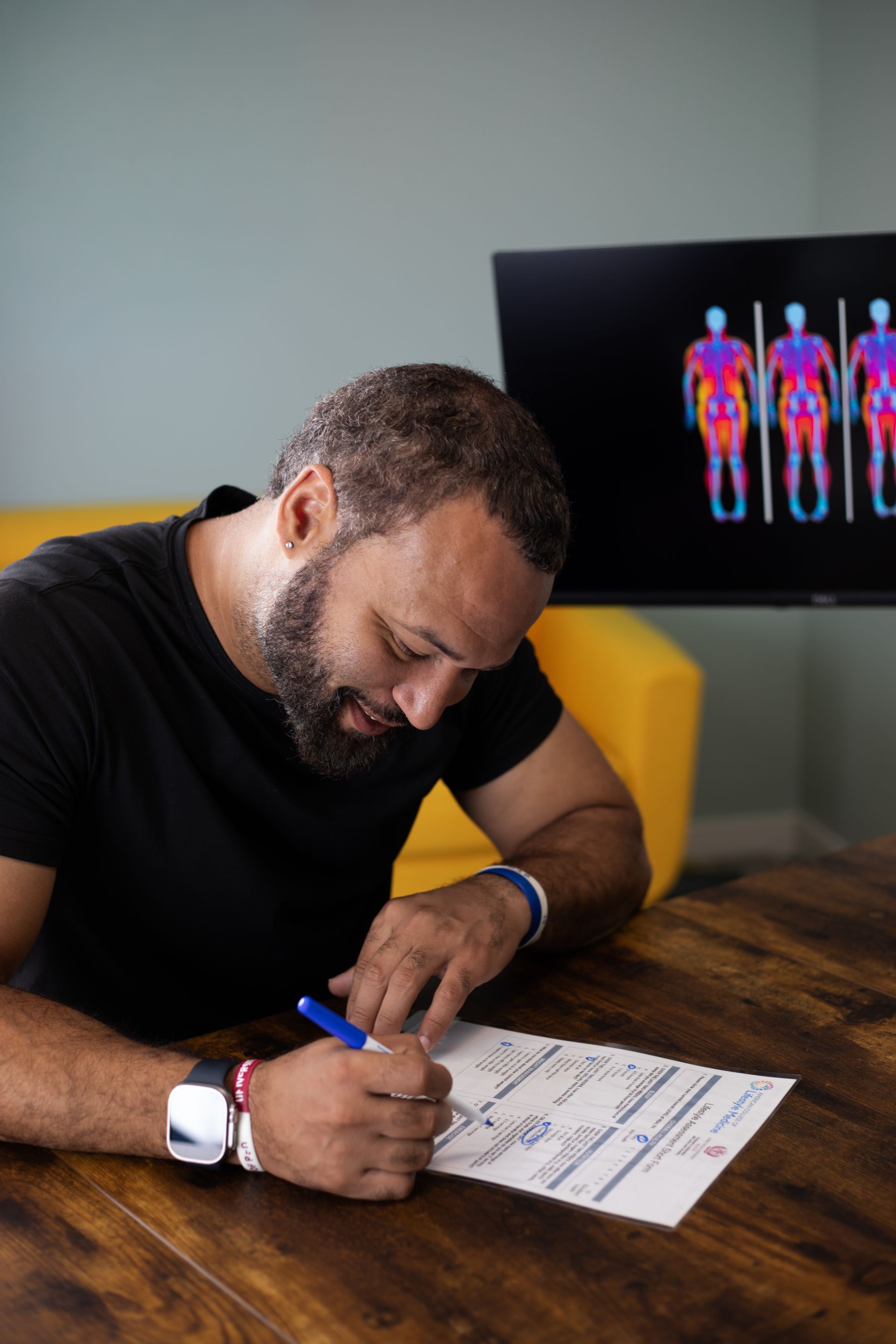Man writing at a table, looking at paper. Thermogram display on TV in background.