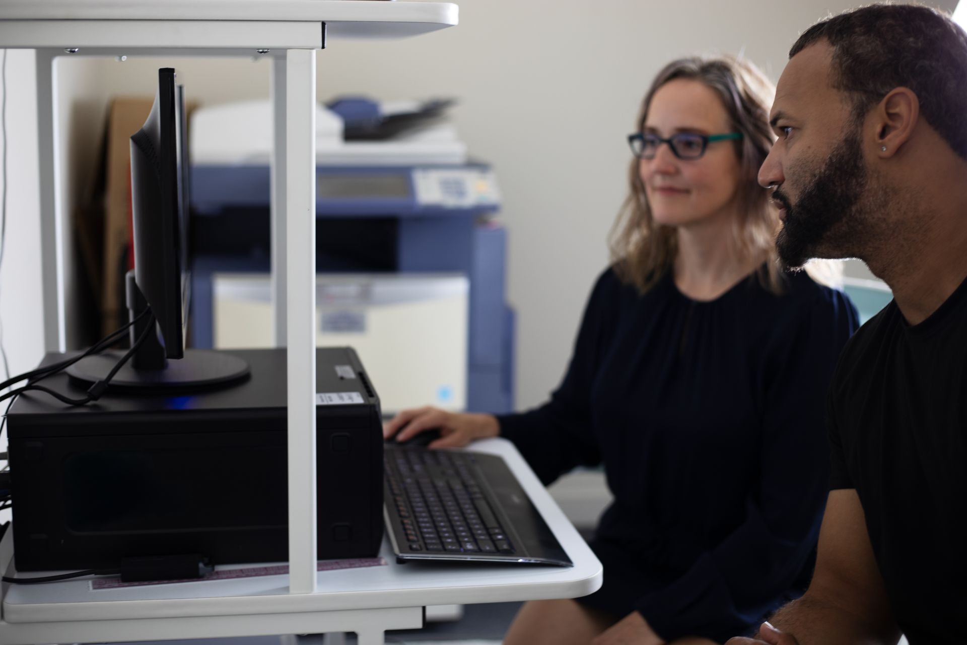 Two people looking at a computer monitor. One person is typing on the keyboard. A printer is in the background.