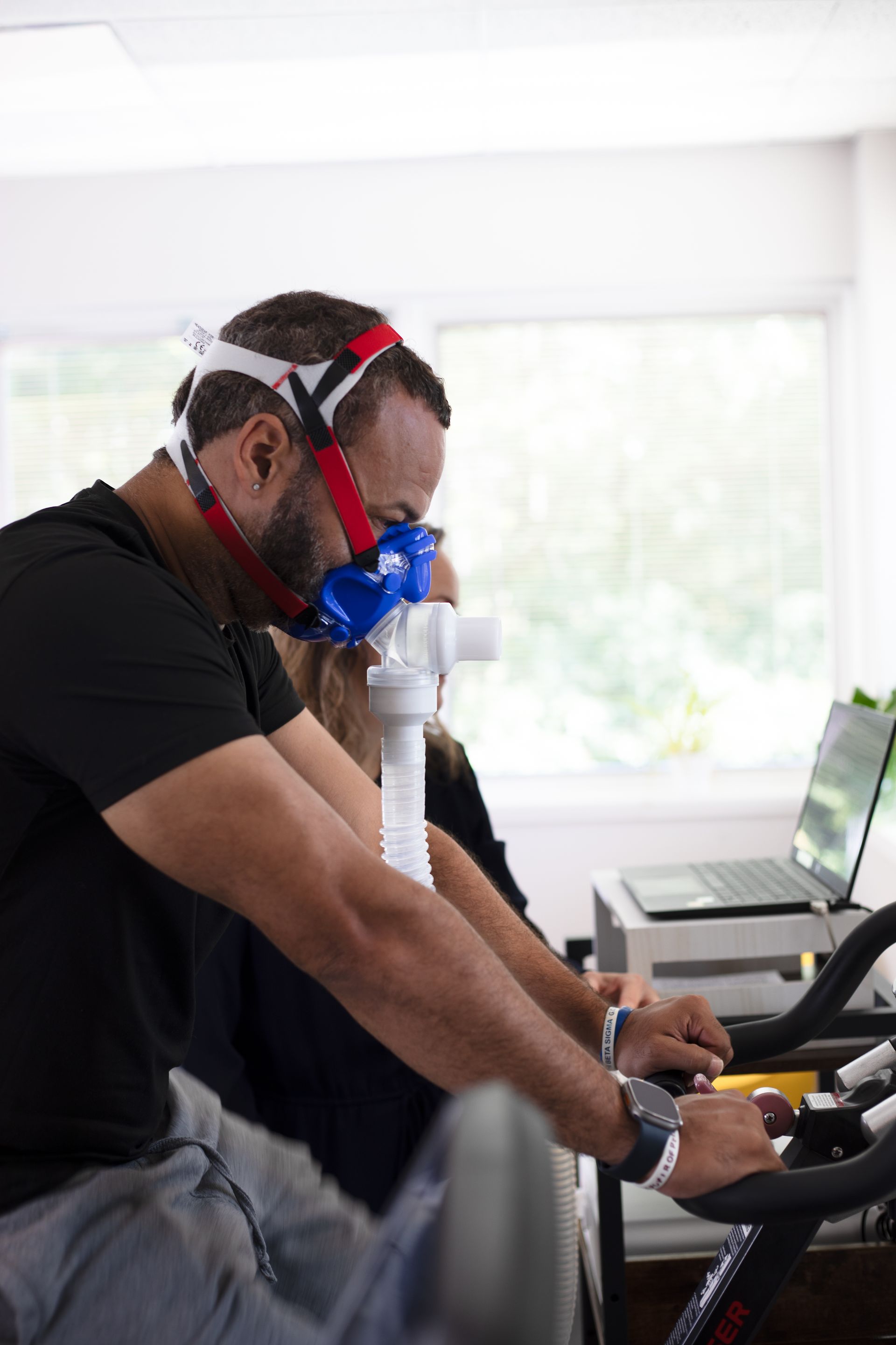 Man wearing breathing mask, cycling indoors while test is performed. White tube connected. Laptop nearby.