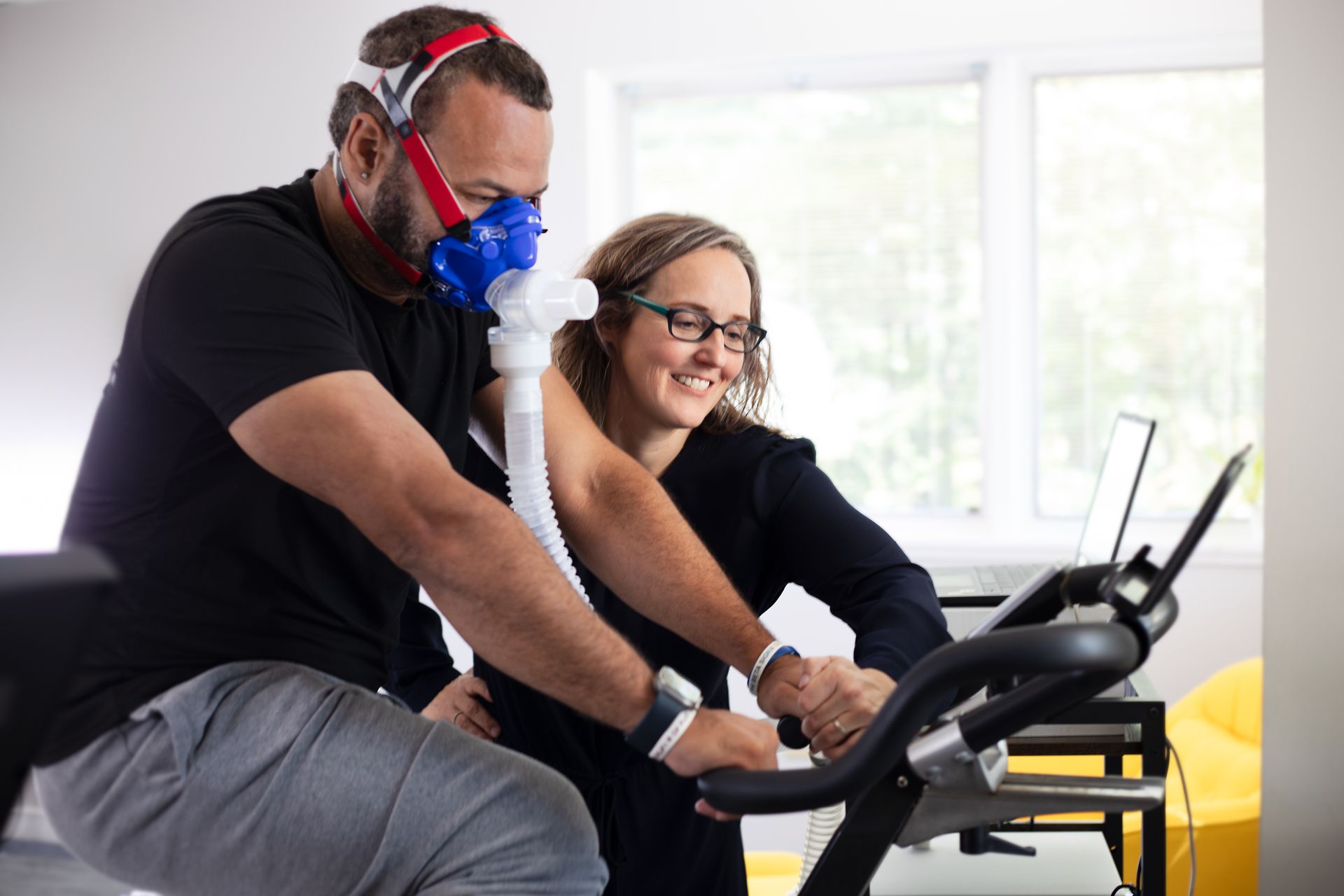 Man on exercise bike wearing mask, being assisted by woman looking at a computer; indoor setting.