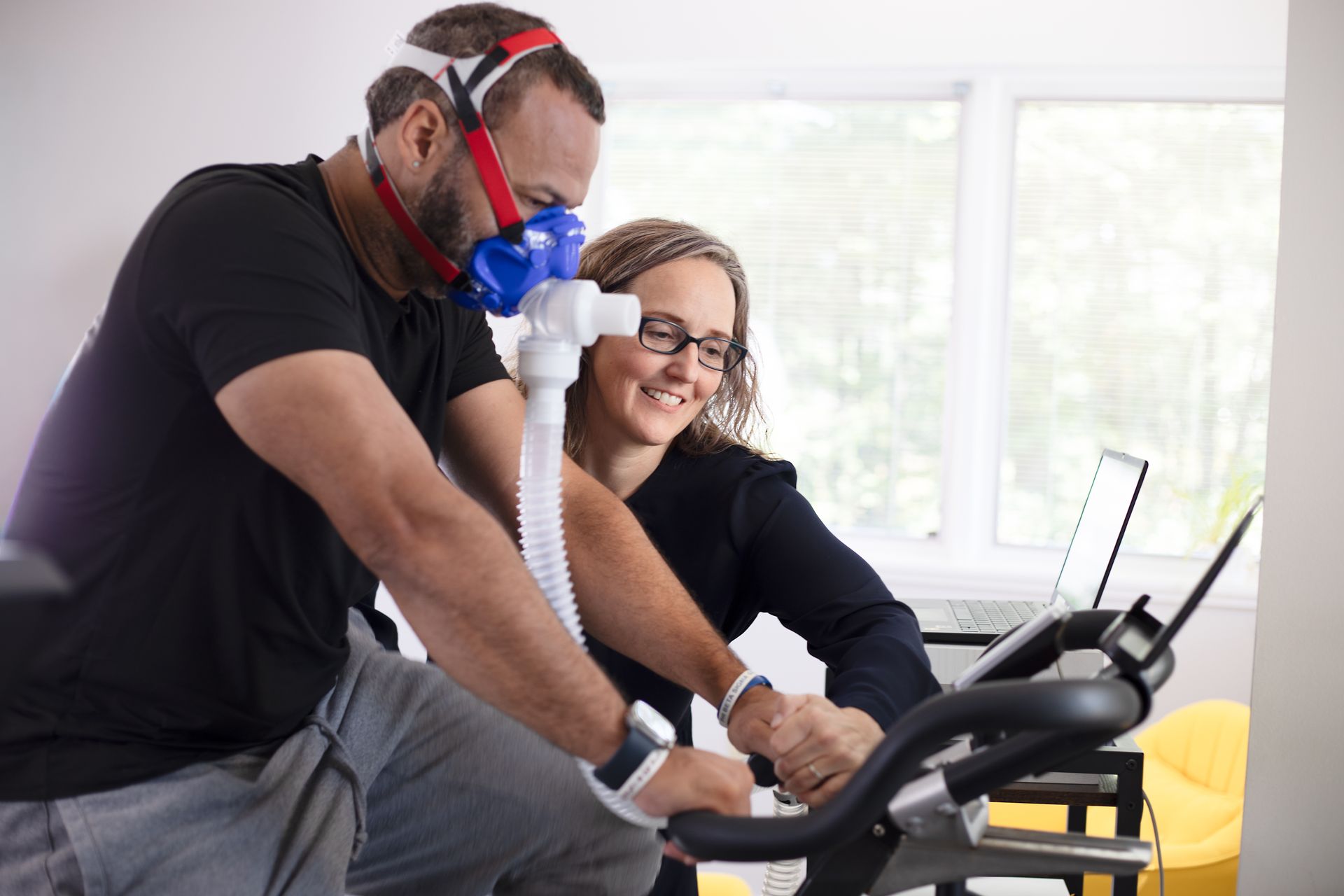 Man on exercise bike with mask, being monitored by a woman with a laptop. In a room.