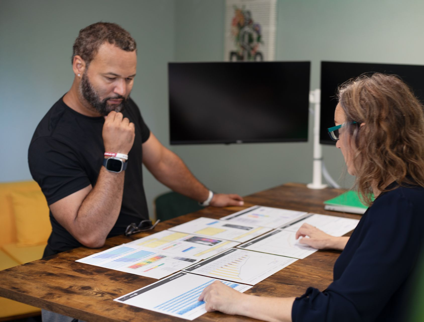 Two people reviewing documents at a table; one with a beard, looking thoughtful. Computer monitors in background.
