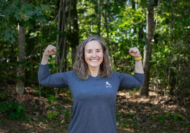 Woman flexing muscles, smiling in front of a forest. She wears a grey long-sleeve shirt.