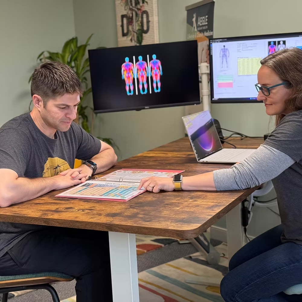 A person reviews a chart with a healthcare provider at a desk. Two monitors display data.