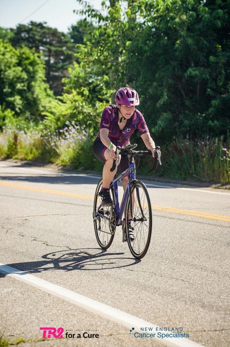 Cyclist in purple gear riding on a sunlit road with trees in the background