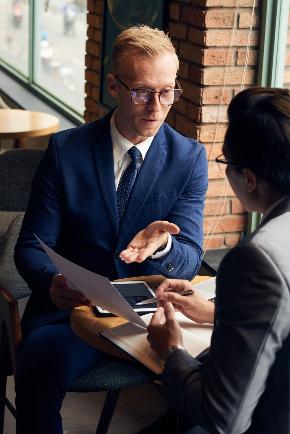Two people in suits discussing documents at a table by a window, in a coffee shop-like setting.
