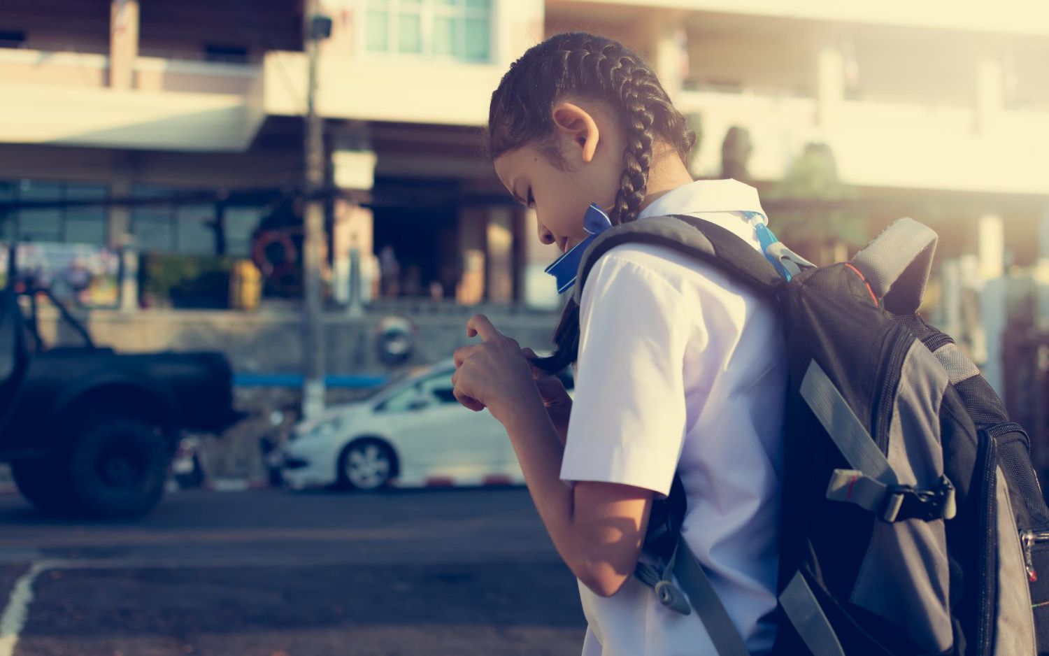 Girl in school uniform, braids, looking at phone, backpack, near street with cars.
