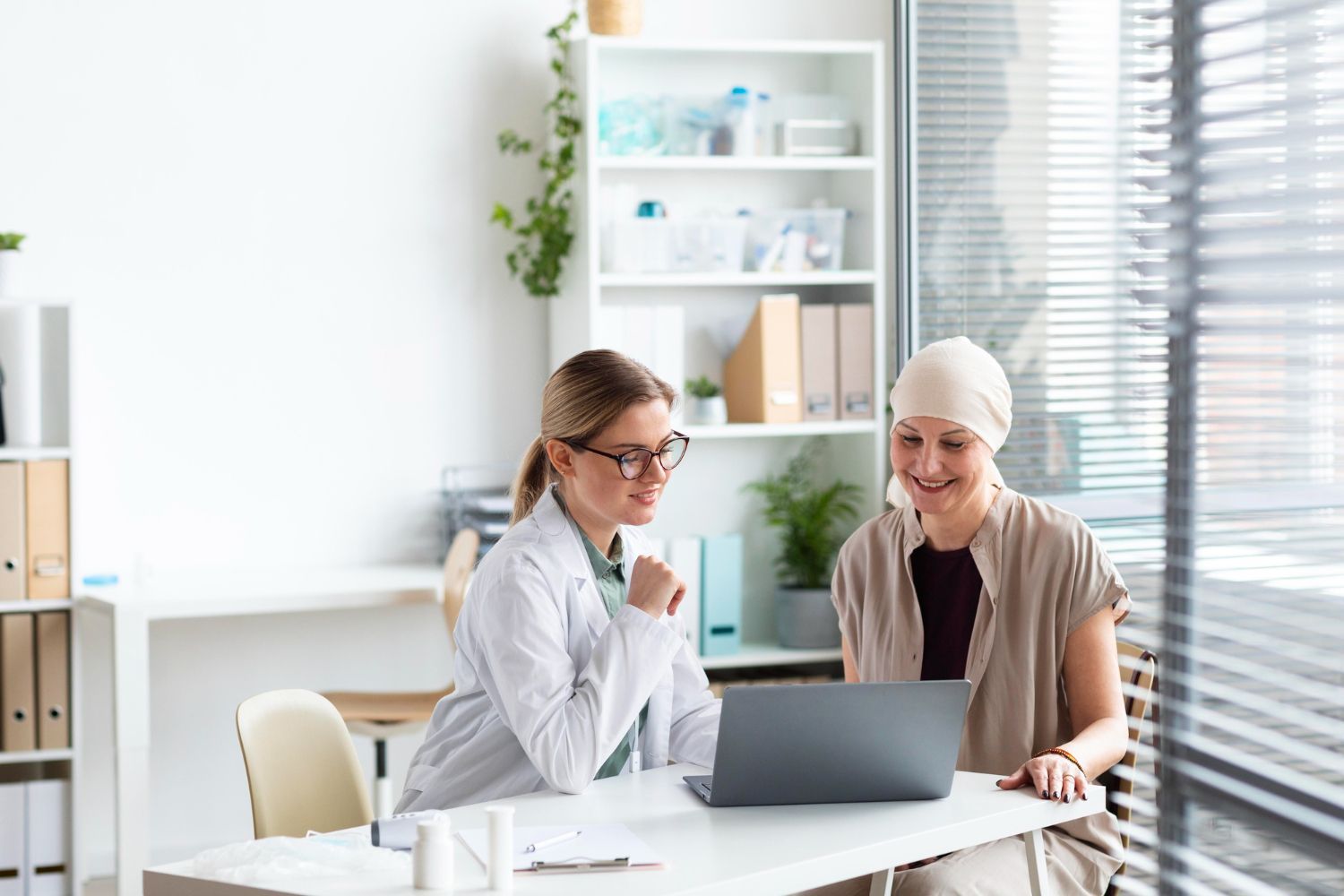 Doctor and patient looking at a laptop in a bright office. Patient wears headscarf, possibly undergoing cancer treatment.