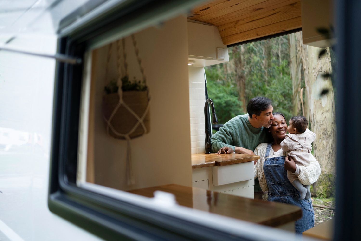 Family smiles together inside a camper, kissing baby. Window frame foreground. Outdoors setting.