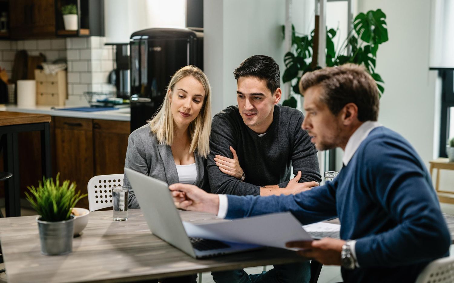 A financial advisor reviews a laptop with a couple in a modern kitchen.