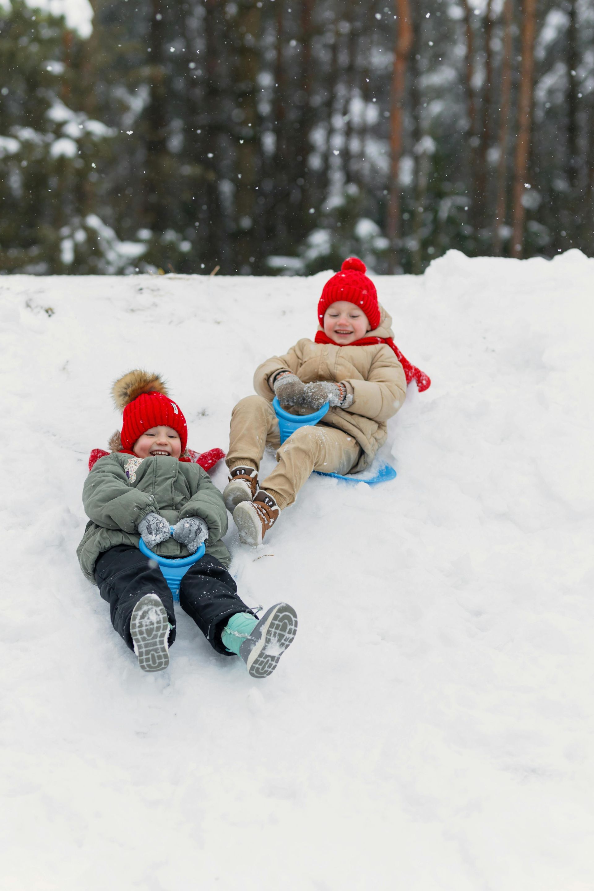 Two children in red hats and coats sledding down a snowy hill, smiling.