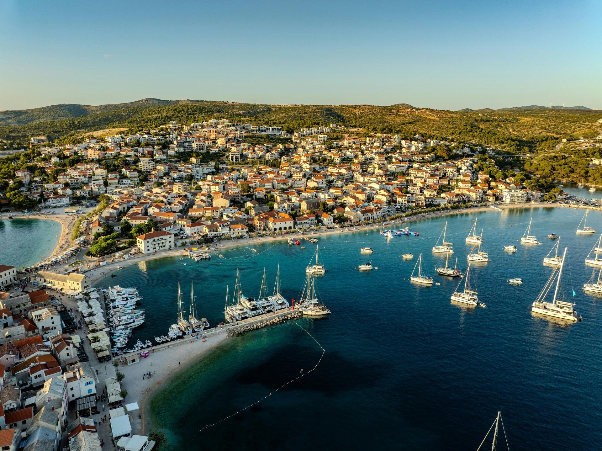Coastal town with harbor filled with sailboats and a beach, buildings on a hillside under a blue sky.