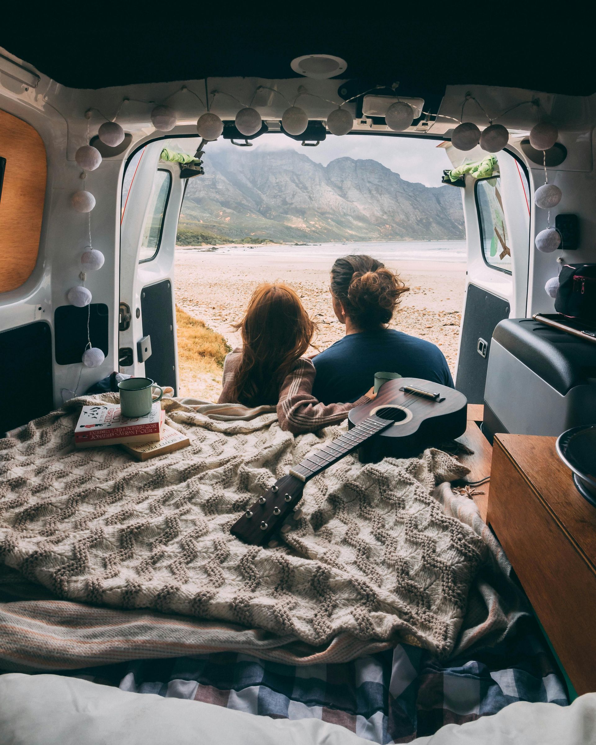 Couple in a van looking out at a beach, with a guitar. Beige blanket, string lights.