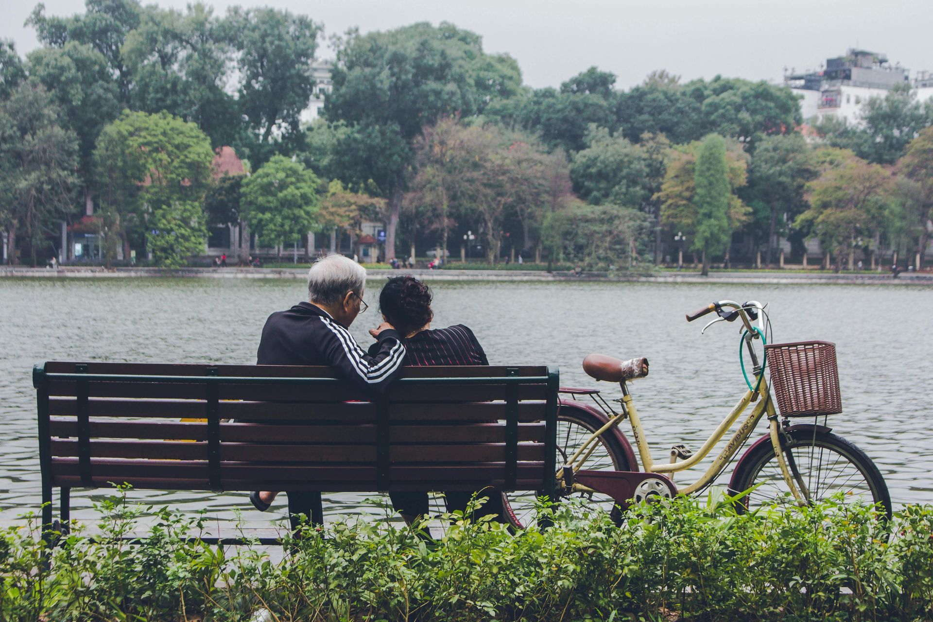 Elderly couple on park bench, bike next to them; overlooking a lake with trees in the background.