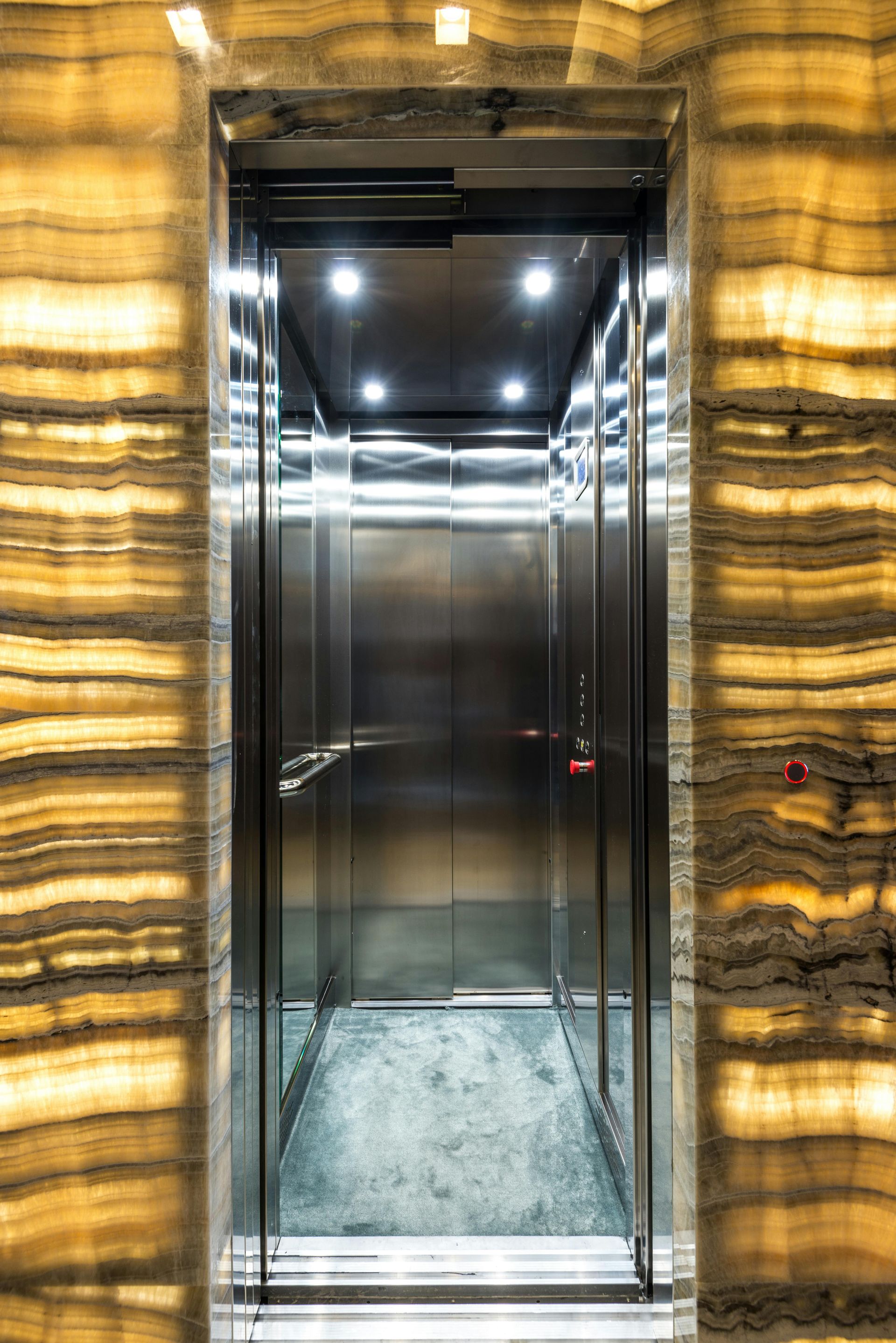Elevator with stainless steel interior; flanked by amber-colored, layered wall panels.