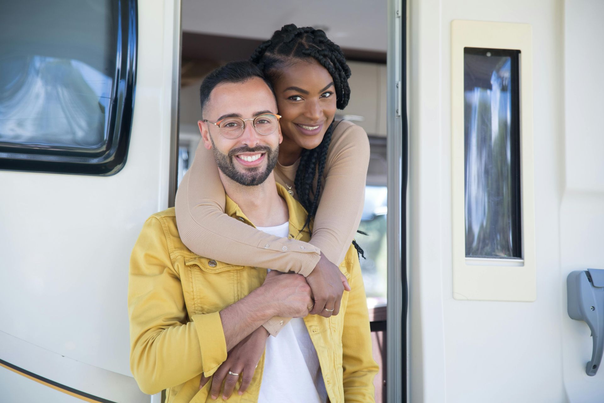 Smiling couple embracing in front of a white camper. The woman has her arms around the man, both looking at the camera.