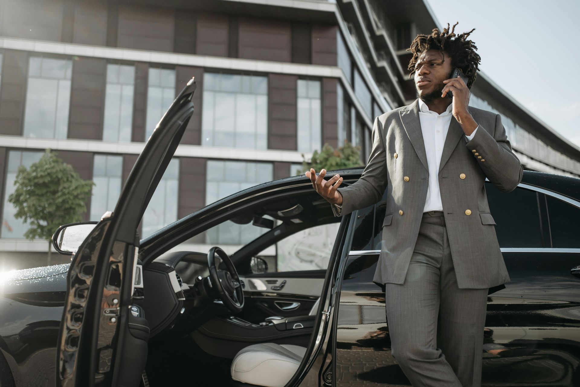 A stylish Black man in a gray suit leans against a black car while on a phone call, near a modern building.