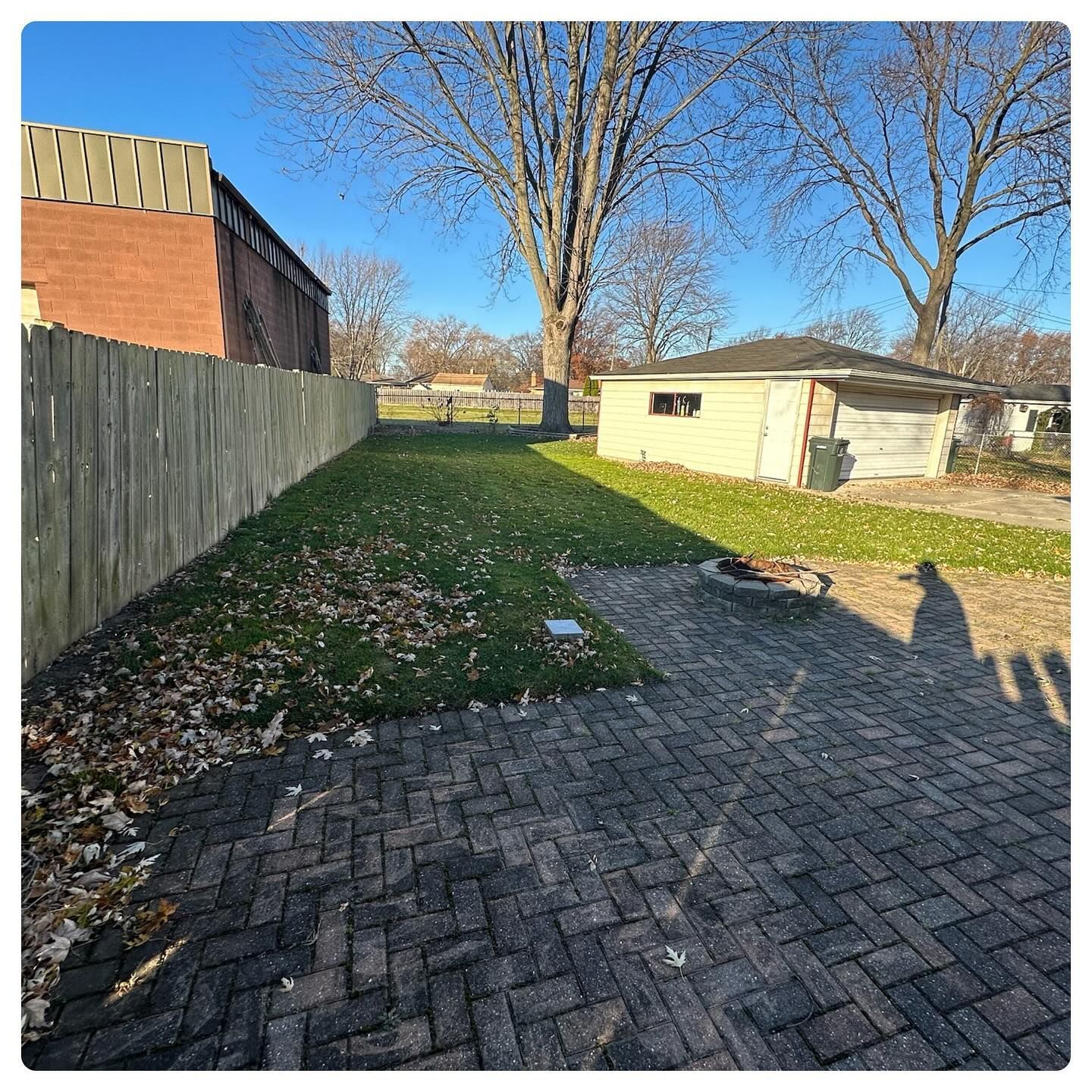 Backyard with brick patio, wooden fence, grass, and trees beside a small shed in sunlight