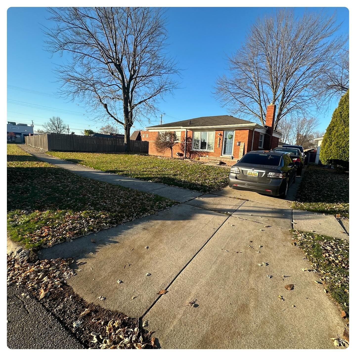 Suburban brick house with driveway, black SUV, leaf-covered lawn, and bare trees on a sunny day