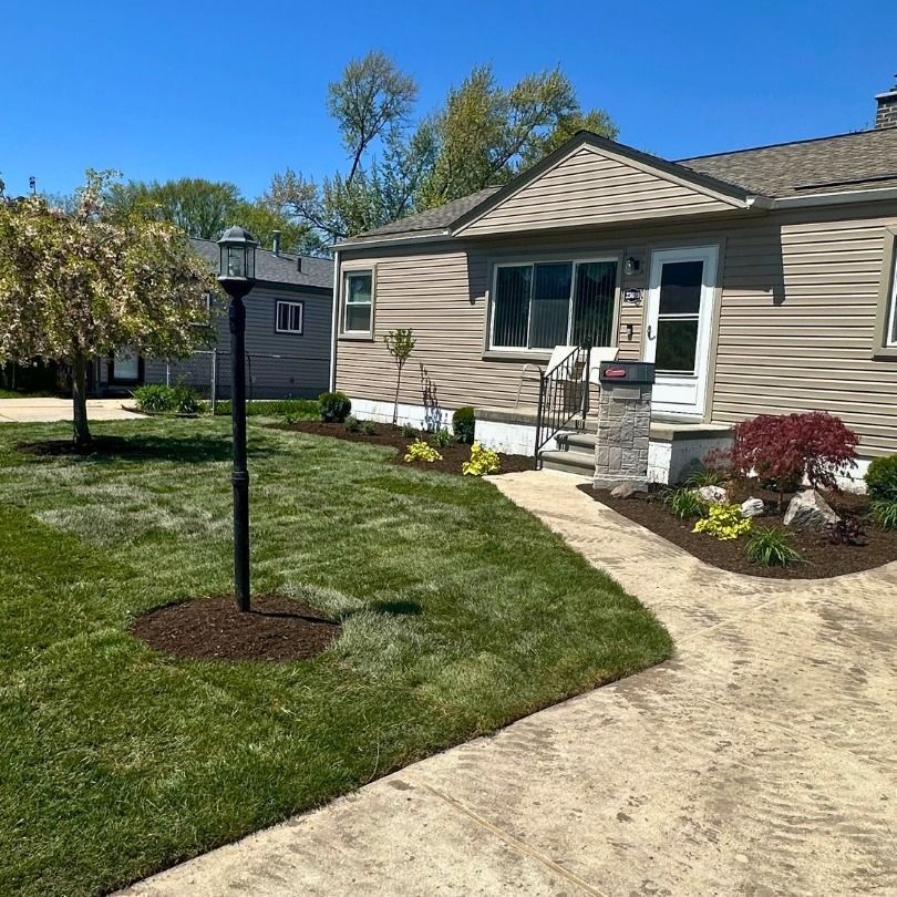Front yard with a beige house, curved walkway, trimmed lawn, and small landscaped flower beds under a blue sky