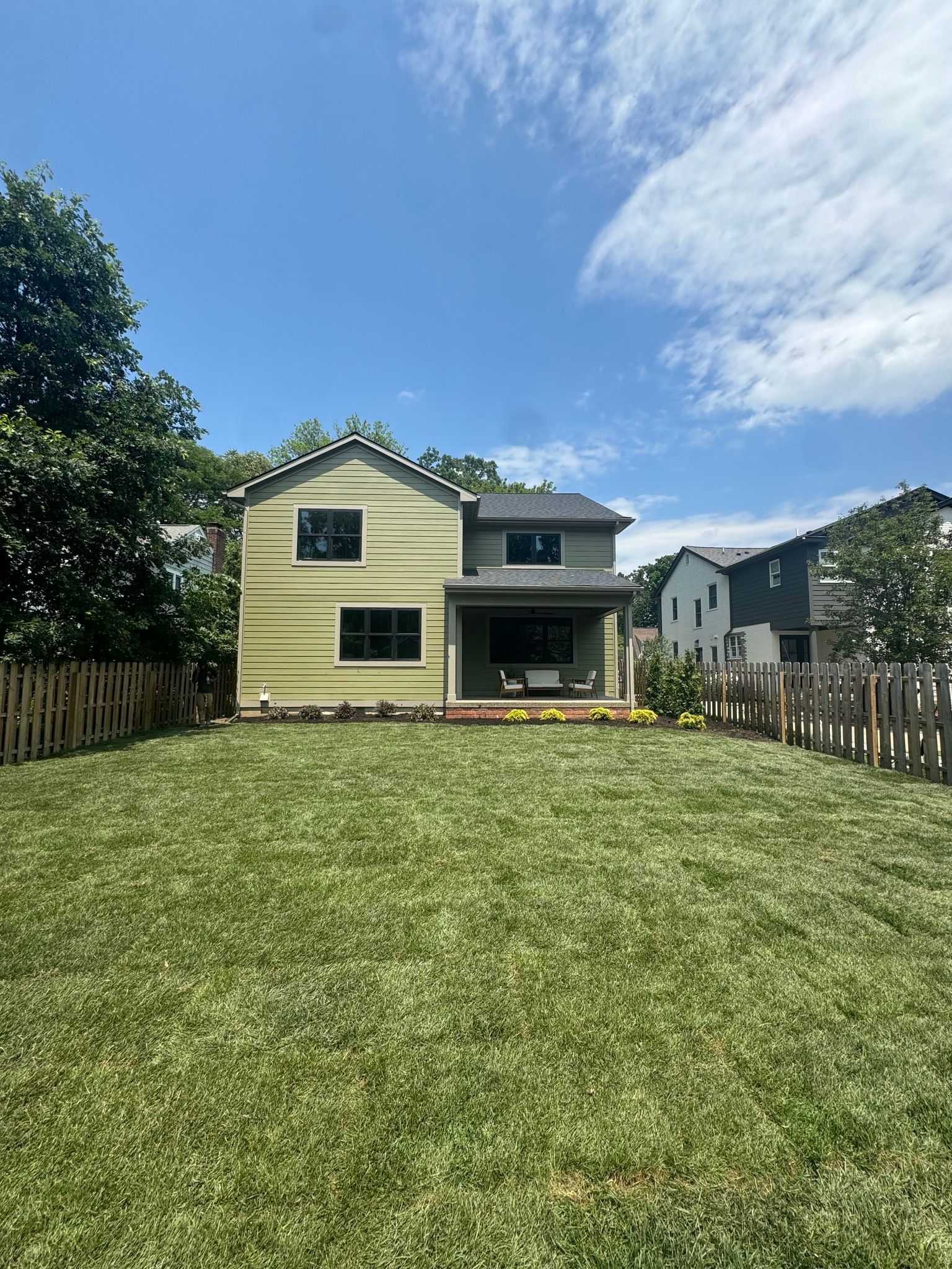 Backyard with a pale yellow house, green lawn, and blue sky with clouds