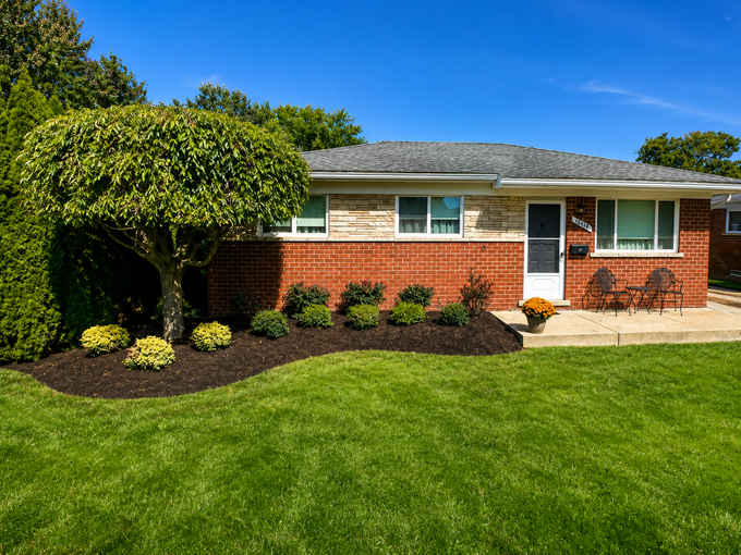 Single-story brick house with manicured lawn, shrubs, and a trimmed tree under a blue sky