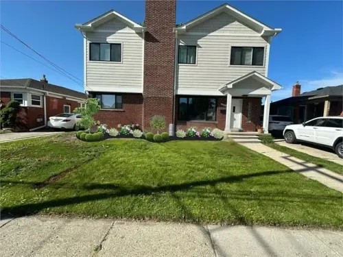 Two-story duplex with brick center, green lawn, and a white car in the driveway on a sunny day