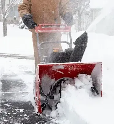 Person using a red snowblower to clear a snowy driveway in heavy snowfall