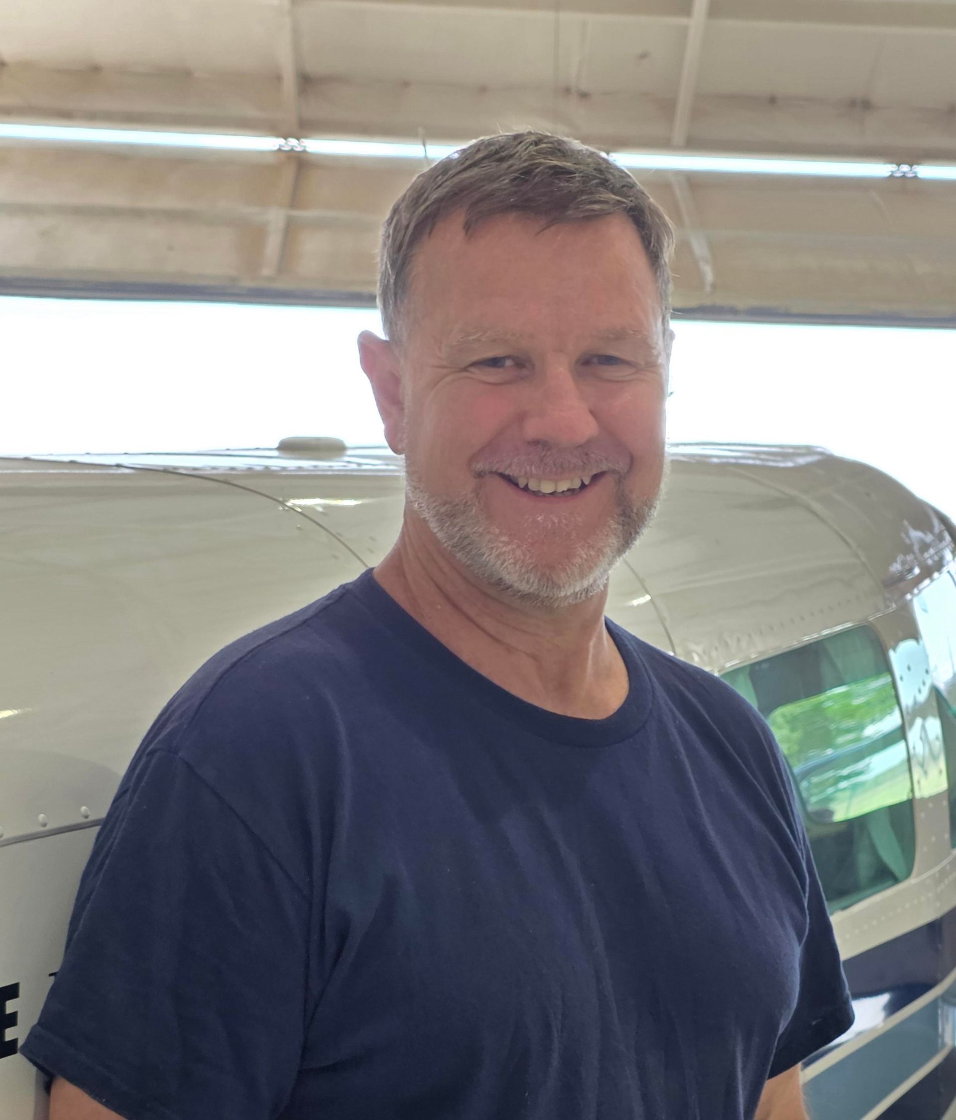 Smiling man in a navy T-shirt standing in a garage beside a covered boat