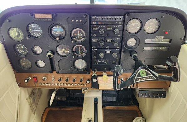 Small aircraft cockpit with analog gauges, radio stack, wooden panel, and yoke.