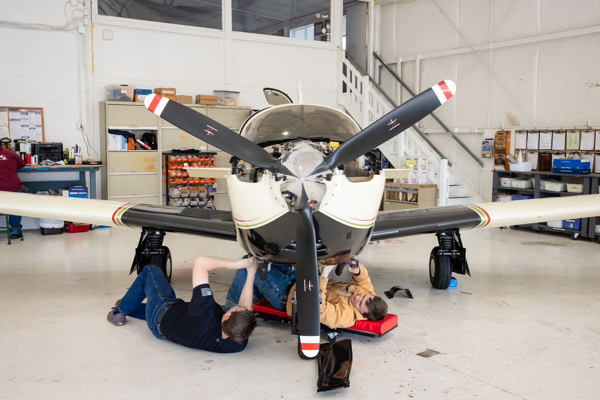 Mechanic working beneath a small airplane in a hangar, with the propeller centered in front.