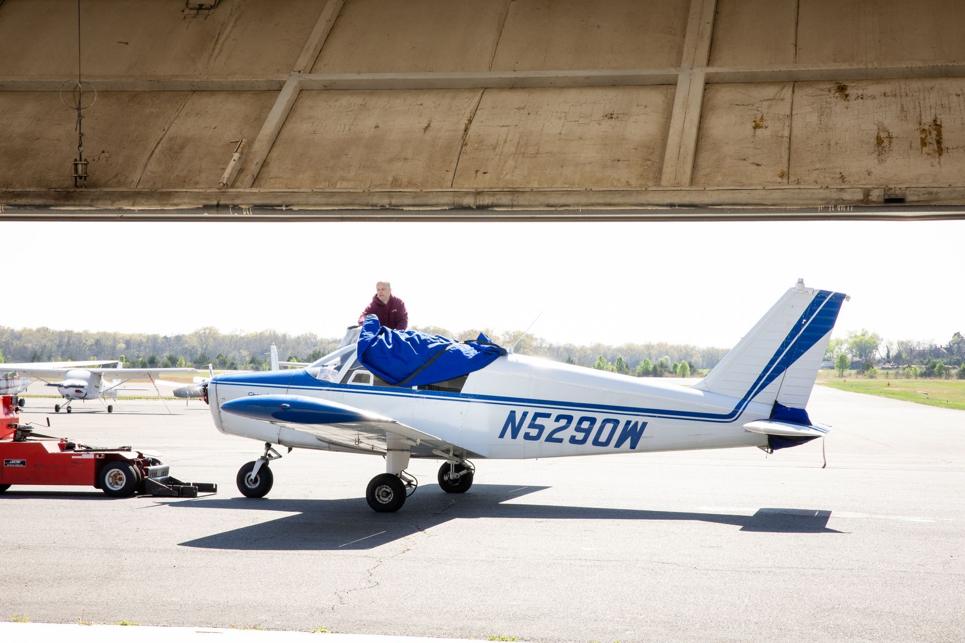 Blue-and-white small airplane on tarmac under a hangar, with a person in the cockpit and a tow vehicle nearby