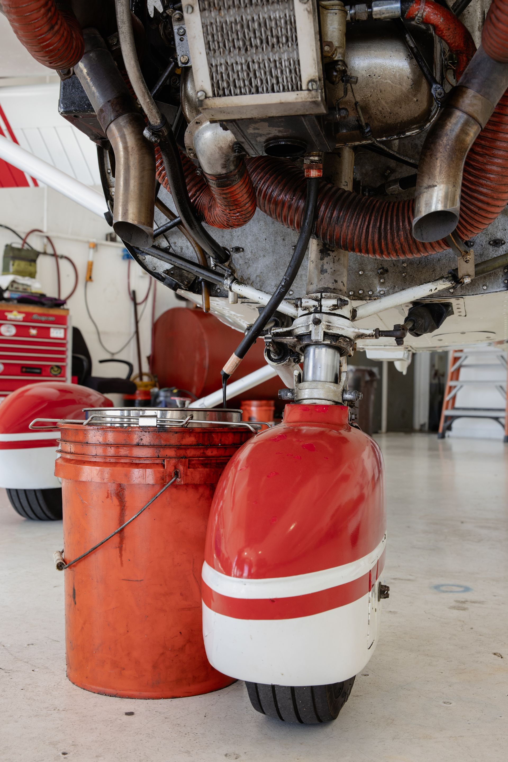 Red-and-white fuel tank beneath an aircraft engine in a hangar, with an orange bucket nearby
