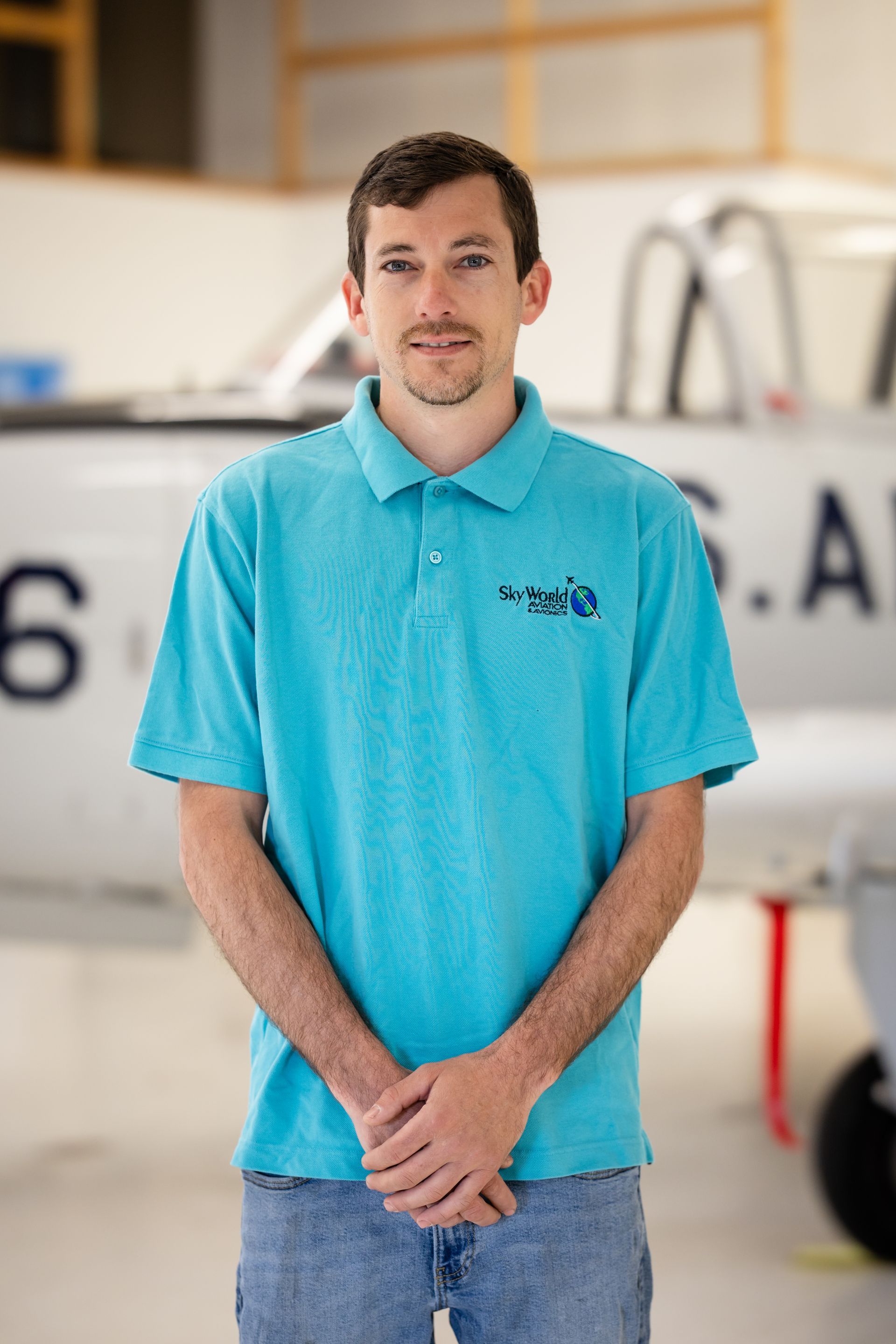 Man standing in a blue polo shirt in front of a small airplane in a hangar
