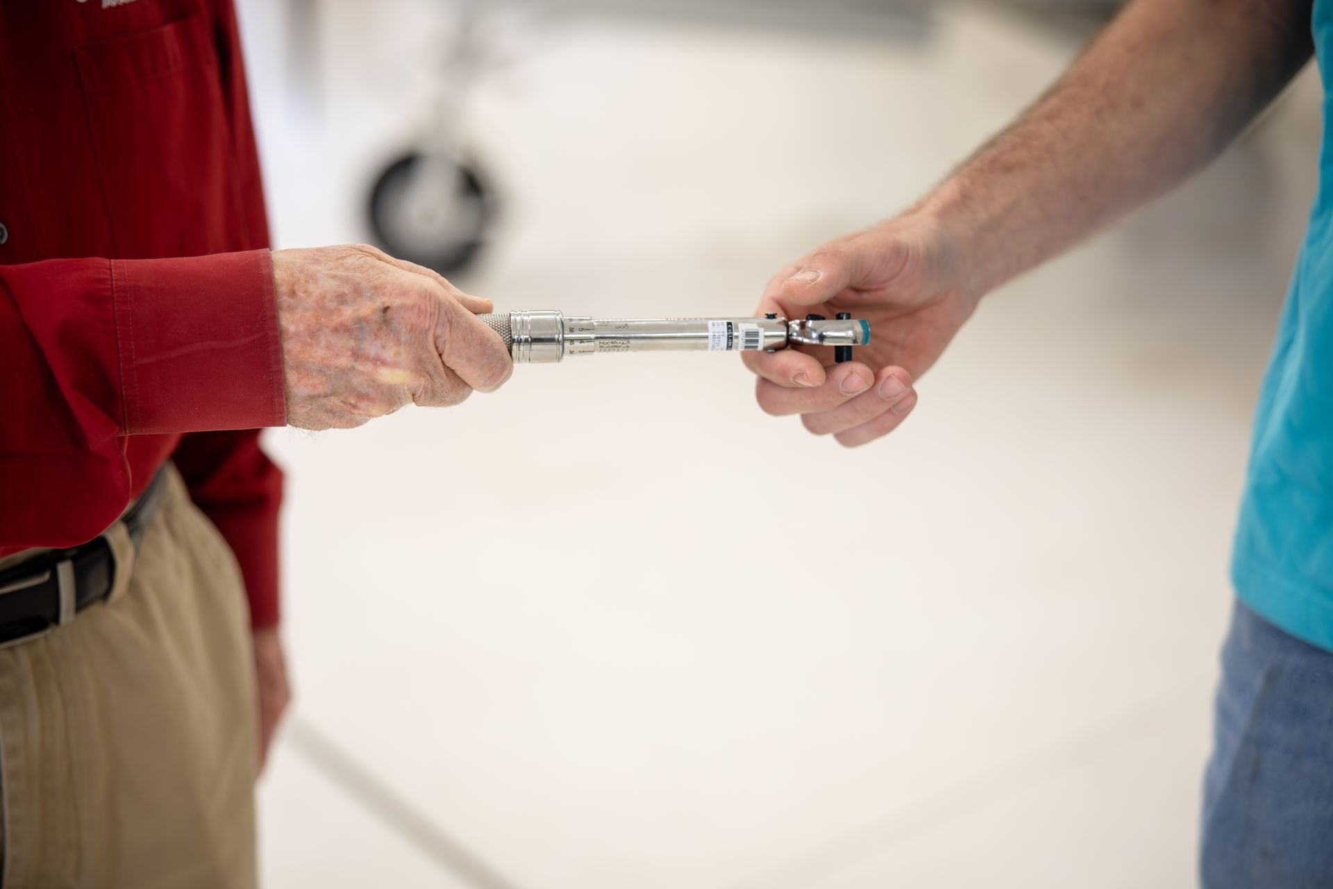 Two people exchange a silver batons-like tool across a white floor, one in red and one in blue.