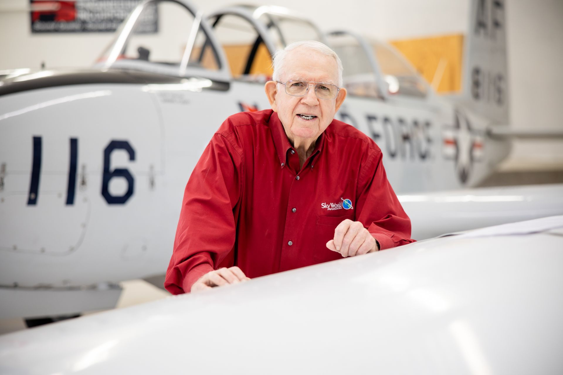 Older man in red shirt beside a small white aircraft in a hangar