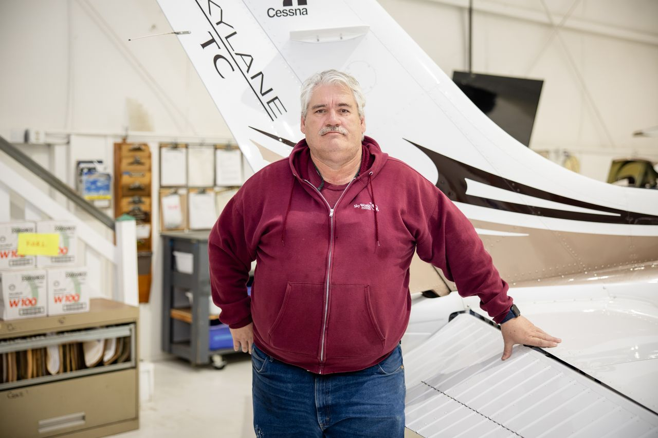 Man standing in a hangar beside a white aircraft, wearing a maroon hoodie and jeans