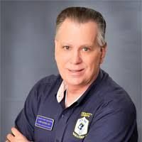 A man in a blue shirt is standing with his arms crossed and smiling.