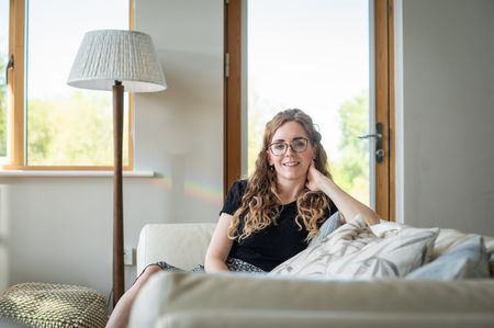 Rachel, sitting on a cream couch, smiles. Beside her is a floor lamp; window behind.