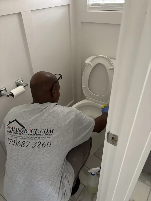 Person kneeling beside a toilet, cleaning a bathroom with a toilet brush and supplies nearby