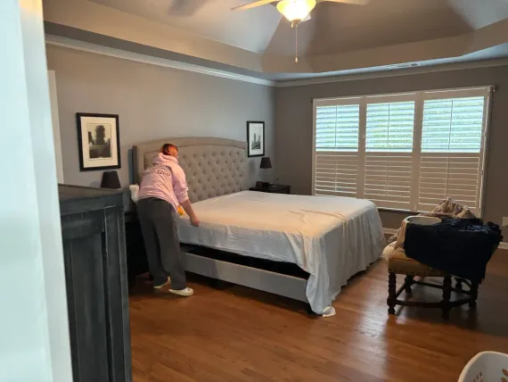 Person making a bed in a gray bedroom with hardwood floors and white shutters.