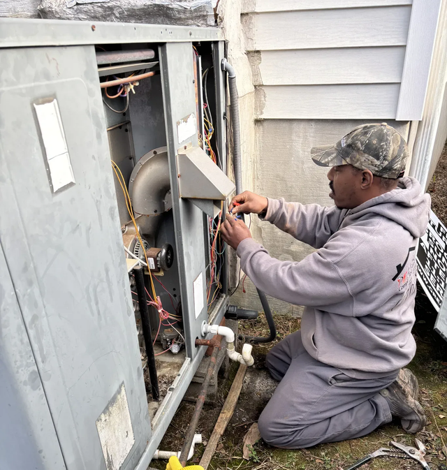 Technician kneels beside an open outdoor HVAC unit, working on wiring with tools.